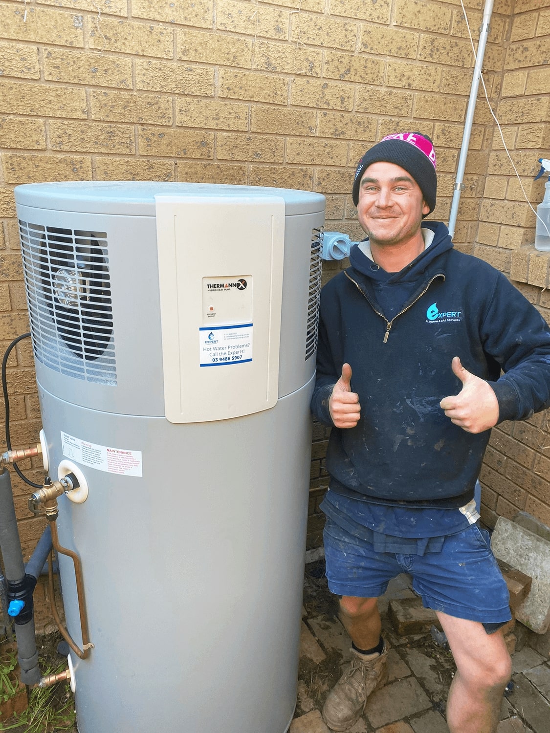 Thermann Hyrbrid Heat Pump System in front of brick wall . A friendly plumber is smiling and giving a thumbs-up