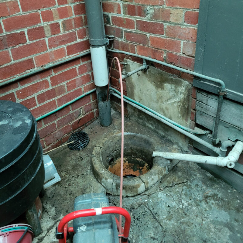 A gully trap with sewer equipment leading into it. There is a red brick wall behind it