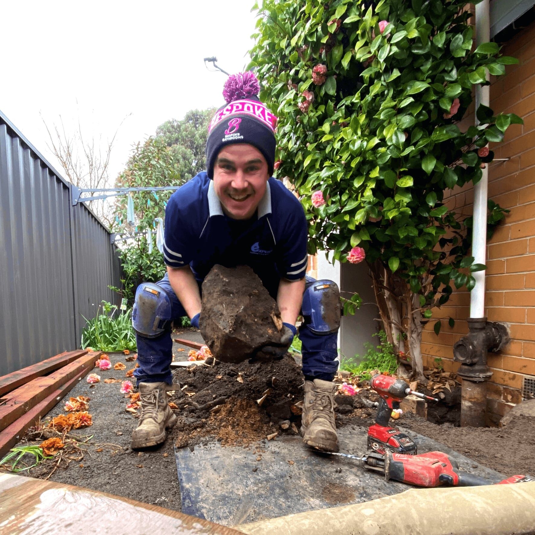 an expert plumber is holding a large rock in his hands. there is a green bush with pink fllowers on the right side. there is a dark grey corrugated fence behind him. The plumber is squatting and has a happy smile on his face.