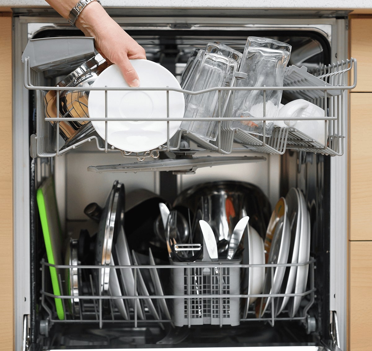 hand putting dishes in a dishwasher. the fishwasher is open and filled with cutlery and culnery