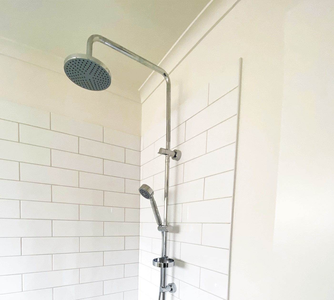 white glossy square tiles on a white wall. a silver chrome twin shower set has a long horizontal pipe and rainfall showerhead and a small european hand shower head attached.